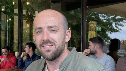 Oliver Long is looking at the camera. He is balding and has a beard and a moustache. He is wearing a green shirt with a white T-shirt underneath. Three men are sitting at tables in a cafe behind him.