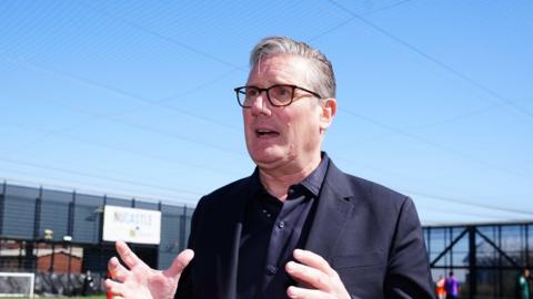 Keir Starmer is gesticulating with both hands held up in front of him while responding to a question. He is wearing a dark navy shirt and suit jacket. The sky is bright blue behind him.