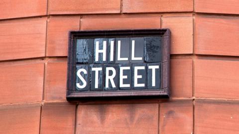 A close-up of a black tiled sign displaying Hill Street in white text. The sign is placed against a terracotta brick wall.
