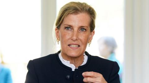 The Duchess is seated at a wooden table in a bright indoor setting, wearing a dark jacket over a light top, gesturing with both hands while speaking. The background is softly blurred, showing two other seated figures and large bright windows.