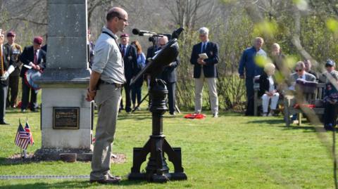 A man stands by a plinth at Markshall, with a microphone surrounded by a crowd commemorating an event.  Some of the crowd are standing, some are sitting down.  The event is held on grass with a stone monument present. Small American and British flags can be seen, sticking out from the ground.