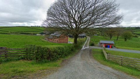 Tan y Dderwen Farm in Betws-yn-Rhos. A house can be seen in the centre behind a large oak tree. A lane road runs in the centre, with green fields either side. Rolling hills can be seen behind the house along with farm sheds. 