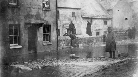 Black and white photo from 1924 - 100 years after the Great Gale - showing people standing around on a flooded street lined with terraced houses
