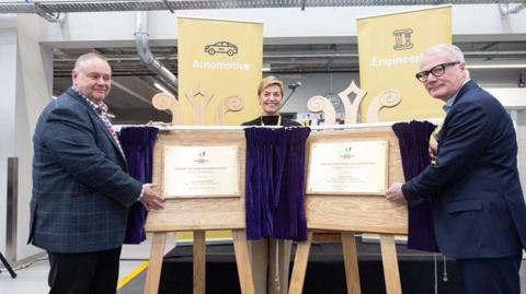 Two men are standing next to wooden boards with commemorative plaques on them, and purple curtains. A woman stands between them, smiling. There are yellow banners in the background