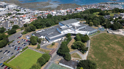 An aerial photograph showing a large building complex set within landscaped grounds. Surrounding the building are car parks filled with vehicles, a green bowling lawn, open playing fields, and a children's playground. Beyond the site lies a densely built coastal town, a marina with moored boats, and a rocky shoreline exposed by low tide, with blue sea stretching across the top of the image.