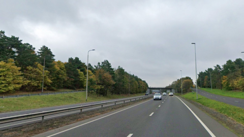 The four lane road has a metal barrier running down the central reservation. The road, which is bordered by trees, has two slip roads coming off of it at an incline on either side. There are half a dozen cars in the distance.