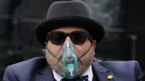 Nicholas Rossi, weaing a blue pin-stripe suit, bow tie and a black bowler hat. He is unshaven and is also wearing sunglasses and an oxygen mask