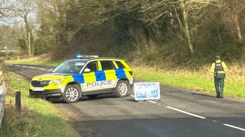 A police car is parked across a country road with a blue police accident sign placed on the road. A police officer is to the right of the road with his back to the camera. In the background we see the road, which is lined with a grass verge and trees, continue into the distance. 