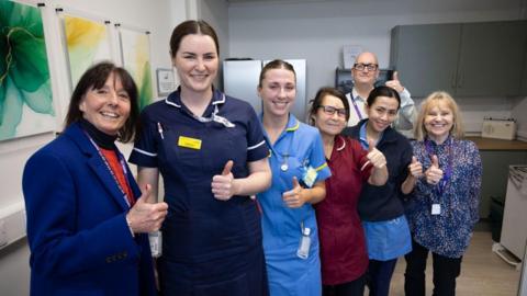 Seven members of staff ( six female and one male) in one of the newly refurbished staff areas. They are all stood smiling at the camera with one of their thumbs up. Four women are wearing traditional hospital scrubs, while two women and one man are wearing traditional office wear. 