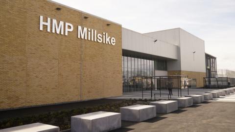 The outside of the jail. Light brown brickwork to the left with the sign HMP Millsike in large white letters, while to the right is a glass-front entrance.