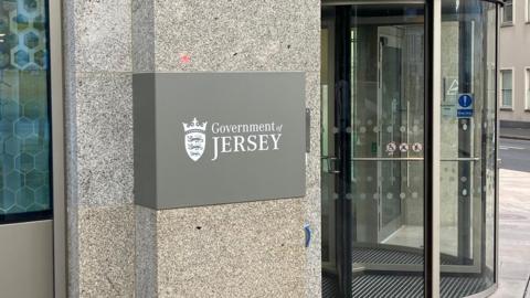 The entrance of the Government of Jersey's building. There is a grey sign with white text, and a revolving door.