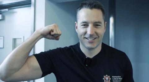 Andy Mitchell smiles for the camera flexing his right bicep. He has short, neat brown hair combed to the side, and wears a dark blue t-shirt with the logo of the Hampshire and Isle of Wight Fire and Rescue Service on the left chest area. 