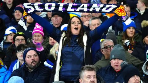Scotland fans in the stands watching a football match. One fan is standing up wearing a Scotland top and holding a Scotland scarf, wile other fans sitting around her have Scotland hats on 