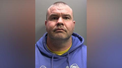 A mugshot of Craig Berry who is staring into the camera with a straight face. He has closely shaved grey and brown hair and is wearing a purple hoodie on top of a bright yellow t-shirt which is just peeking through.