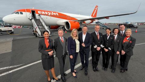 EasyJet pilots and crew stand in a line in front of an orange and white plane which is parked on the tarmac. They are wearing black and orange uniform and smiling brightly at the camera. There is also a man and a woman in the middle, wearing suits.
