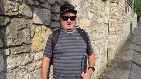 Andy Evans is standing on a pavement in front of a stone wall, wearing a hat and a backpack. In his hand is a white cane, and he is wearing the AI glasses, looking at the camera.