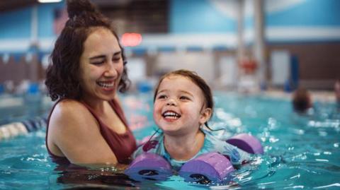 An adult supports a small child wearing purple armbands in an indoor swimming pool as the child practises floating and moving through the water.
