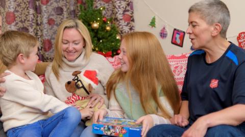 Two children sitting on a couch receiving Christmas presents with their mother in between them and a representative from the Salvation Army next to them.