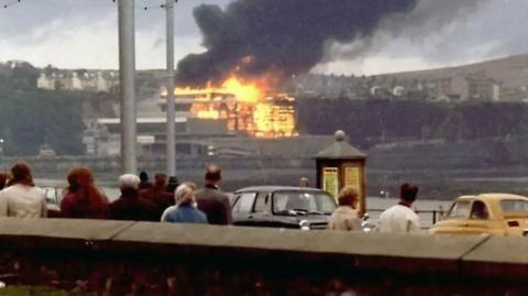 People on Douglas Promenade standing next to cars from the time looking on as the Summerland complex is engulfed in flames across the bay.