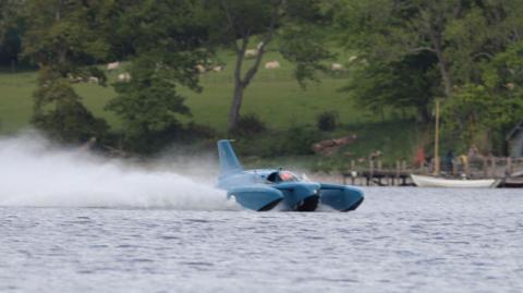 Bluebird K7 "planes" on Coniston Water. The blue craft is throwing up a large spray behind it as it glides on top of the lake's surface. On the far shore, sheep are grazing on an incline.
