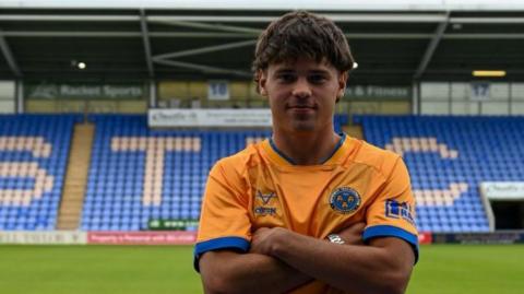 Byron Pendleton folds his arms in a yellow Shrewsbury Town shirt in front of the main stand at Croud Meadow.