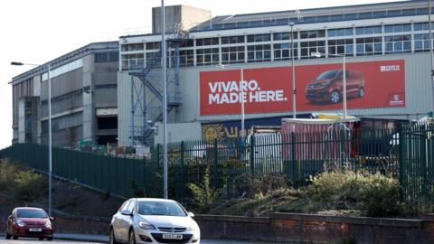 The back of the Vauxhall van plant in Luton, with a huge red poster on the side saying VIVARO MADE HERE and a photo of a red van. Two large industrial buildings stand side by side, with a blue fire escape at the back. Flanked by a tall green metal fence and a road, two cars drive past.