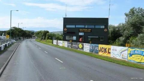 A road with a large black building on the right hand side, barriers are up along the road with different sponsors. 