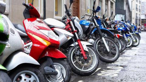 A row of motorcycles - some coloured red or blue - parked on a wet street.  