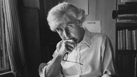 Ronald Blythe pictured in black and white. He is sitting a desk with books open. He is holding his glasses and looking into the camera. 