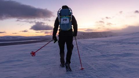 Alex walking across the ice, his back is to the camera, he carries red walking poles and a blue rucksack on his back with yellow straps