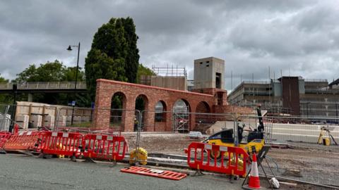 A construction site shows a red brick archway structure under development with scaffolding and safety barriers surrounding it. In the background, a modern building and a pedestrian bridge are visible under a cloudy sky. 