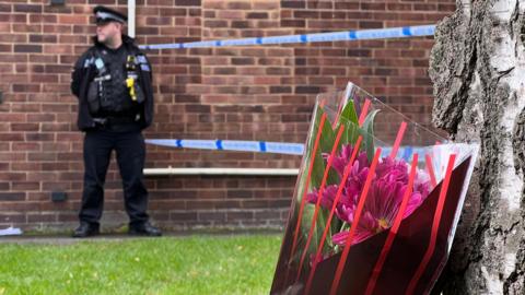 A bouquet of flowers left by a tree. In the photo there is also a police officer stood by a police cordon.