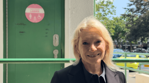 Senator Elaine Millar wearing a dark jacket, standing outside a public toilet entrance, with a green accessible restroom door visible in the background.
