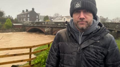 Egremont town councillor David Wright who stands in front of a swollen river under an old bridge. He is wearing a black coat, black hat and grey hoodie. It is raining heavily. 