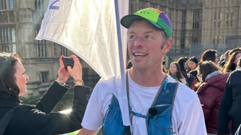 The image shows a male runner standing on a busy street near the Palace of Westminster in London. The runner is holding a large white flag attached to a pole. The runner is wearing a white T‑shirt, a colourful cap and a blue running vest with pockets and straps.