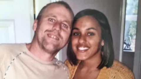 A smiling man and woman looking at the camera. The woman has dark shoulder length hair and is wearing a knitted mustard coloured top. She is stood closely next to a man, with their heads touching. He has short hair and facial stubble and is wearing a sand coloured t-shirt. 