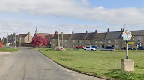 High Street in Woodford is mostly residential properties. There is a patch of grass to the right with a sign for Woodford and a war memorial in the background. 