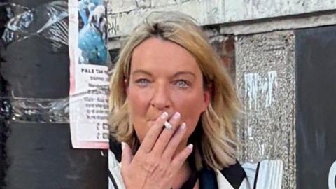 Louise Gallagher, photographed leaving court. She is seen emerging from an alley and is smoking a cigarette. She has blonde shoulder-length hair.
