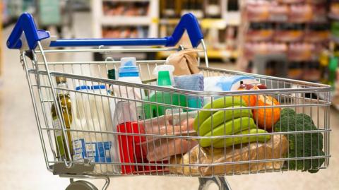 A shopping trolley in the middle of a supermarket aisle. It is filled with grocery items, like milk and bananas. Surrounding the trolley are shelves, but the contents are out of focus.