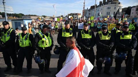 A man with a St George flag walk in front of a line of police officers, with anti-racism protesters a few metres behind 