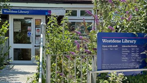 The image shows the outside of Westdene Library, with lavender planted outside a blue sign on the right, which has been placed behind a metal railing.
On the left-hand-side, the entrance can be seen, with another blue sign above the door reading "Westdene Library".