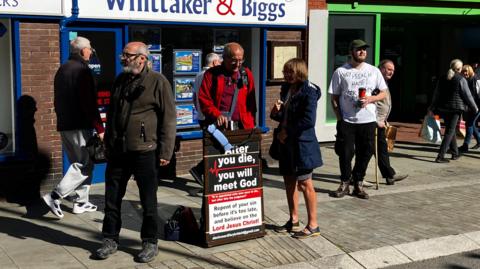 Three preachers stood round a sign on a high street whilst another person stand next to them with a speaker under there arm.