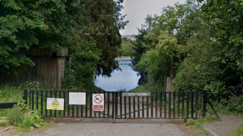 A gated gravel path leading to a large body of water surrounded by trees