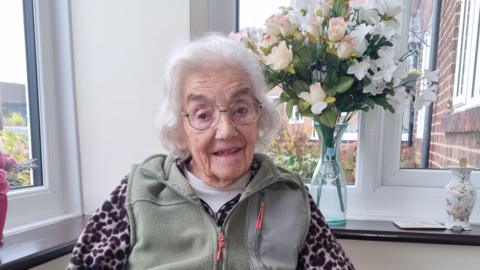 Christina Hartnell is sitting in a conservatory with a large vase of flowers behind her.