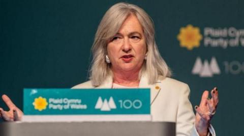 Liz Saville Roberts giving a Plaid Cymru conference speech. She is standing at a lectern holding her hands in the air just below shoulder height. A turquoise sign on the lectern reads Plaid Cymru/Party of Wales 100 in white lettering, beside the party's yellow flower and three white triangles known as the three peaks symbol representing the party's main aims. 