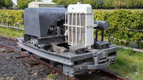 A locomotive engine with a grey base and white and grey machinery on top. It has been placed on a set of rails near a grassy border and a hedge.