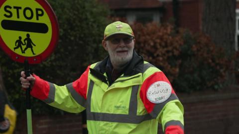 Lollipop man John Astley holds up a lollipop sign bearing the words 'stop' with a symbol of children crossing a road. He is smiling and wearing a high-vis jacket and hat while stood by a crossing. 