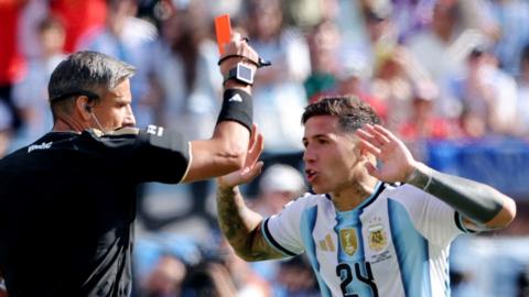 A referee holds up a red card to Argentina's Fernandez who is holding hands up in the air