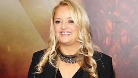 Lucy Davis smiling in a black jacket and necklace on a red carpet