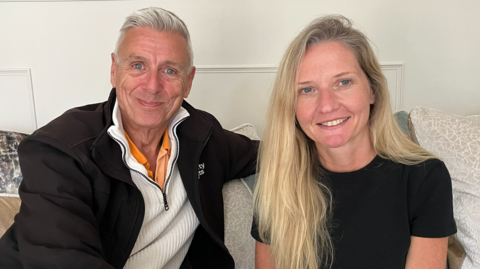 Paul and Lindsay Stone are sitting on a sofa smiling. He has an arm around her and has short grey hair and blue eyes. She has long, blonde hair and blue eyes.